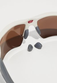 Sonnenbrille mit einem weißen Rahmen, braunen Gläsern und grauen Gummidetails. Drei graue Gumminasenpolster liegen daneben.
