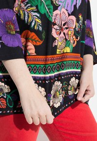 Kurzärmeliges schwarzes Oberteil mit bunten Blumenmustern in Pink, Lila und Grün, kombiniert mit roten Hosen. Das Material wirkt weich und dehnbar.
