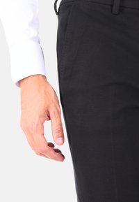 Pantaloni neri texturizzati con una mano posizionata vicino alla tasca. La manica della camicia è bianca, in contrasto con i pantaloni scuri.