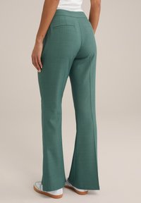 Gröna utsvängda byxor med en slät yta, med en bakficka, bärs med vita sneakers och visar en enkel, skräddarsydd design.