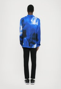 Camicia di seta blu con motivi astratti, maniche lunghe e vestibilità comoda, abbinata a pantaloni neri e scarpe da ginnastica nere.