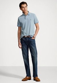Light blue polo shirt with a collar, paired with dark blue jeans. The model wears brown loafers and a brown belt, posing against a neutral background.