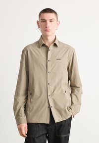 Camicia beige con bottoni, maniche lunghe, logo sul petto, due tasche laterali e bottoni grigi, realizzata in tessuto leggero e testurizzato.