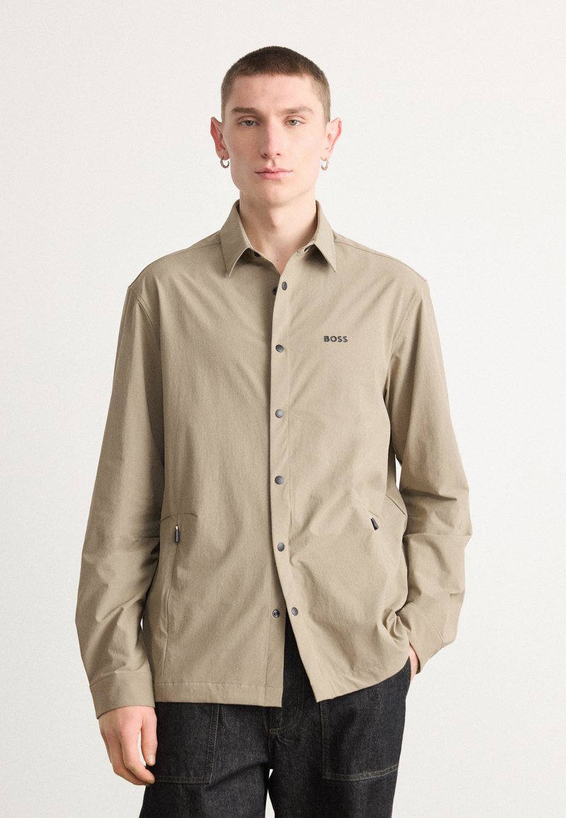 Camicia beige con bottoni, maniche lunghe, logo sul petto, due tasche laterali e bottoni grigi, realizzata in tessuto leggero e testurizzato.