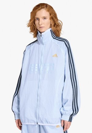 Persona con capelli rossi che indossa una giacca Adidas azzurro chiaro con zip e strisce nere sulle maniche e pantaloni abbinati su sfondo bianco.