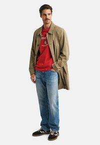 Beiger Trenchcoat, rotes Grafik-T-Shirt, hellblaue Jeans, braune Turnschuhe. Der Mantel hat Knöpfe, das T-Shirt zeigt ein bedrucktes Design mit Schriftzug.