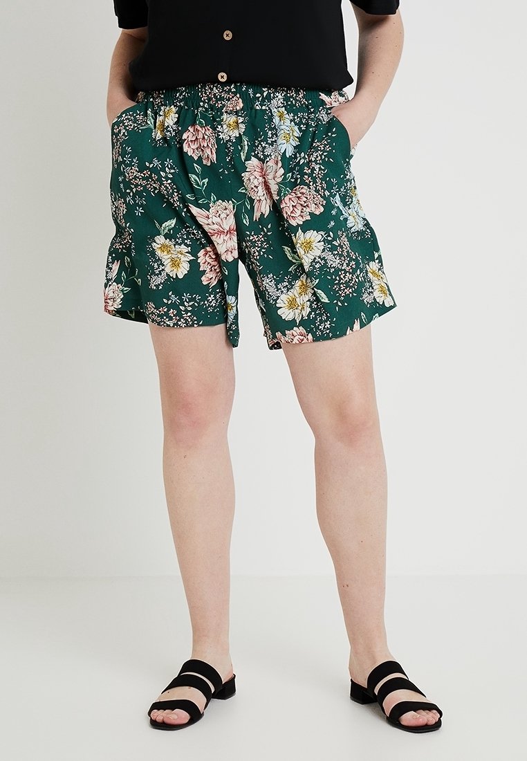 Personne portant un short vert à fleurs, un haut noir et des sandales noires ouvertes à deux brides, debout les mains dans les poches.
