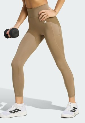 Person iført beige højtaljede leggings og hvide sneakers, der holder en sort håndvægt, stående i en bred atletisk stilling.