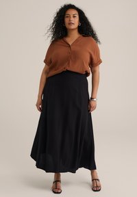 Camicia button-up marrone a maniche corte, con texture, abbinata a una lunga gonna nera. Sandali con sottili cinturini completano l'outfit.