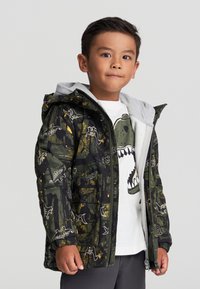 Padded jacket z czarno-zielonym nadrukiem dinozaurów, z szarym kapturem i kieszeniami na zamek. Nosi się na białej koszulce z graphic print.