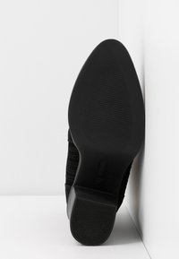Semelle de chaussure noire avec une surface en caoutchouc texturée, présentant de discrètes lignes verticales et un léger talon. Le matériau semble durable et flexible.