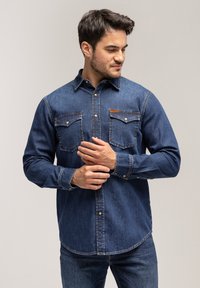 Koszula dżinsowa w ciemnoniebieskim kolorze, z kołnierzykiem zapinanym na guziki, dwoma kieszeniami na piersiach i kontrastowymi detalami szycia. Długie rękawy z mankietami zapinanymi na guziki.
