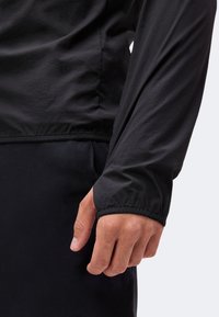 Main et poignet d’une personne portant une chemise noire à manches longues avec un poignet élastique et un pantalon noir sur un fond clair.