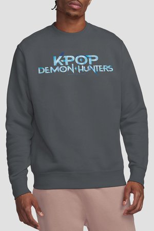 Man wearing dark gray sweatshirt with blue "K-POP DEMON HUNTERS" text, paired with light pink pants against a plain background.