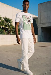 Férfi fehér Bugs Bunny grafikás pólóban, fehér nadrágban és fehér tornacipőben, betonfalak és pálmafák közelében áll a szabadban.