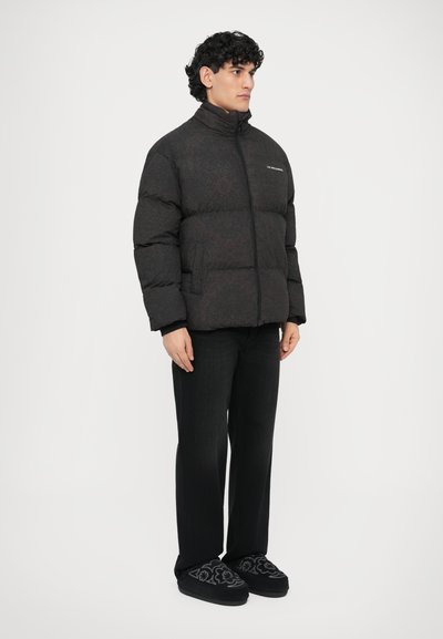 Sort polstret jakke med høj krave, med et diskret mønster. Kombineret med mørke bukser og mønstrede sorte tøfler.