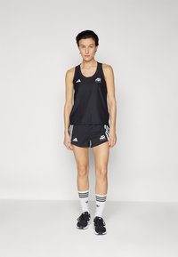 Débardeur de sport noir et shorts avec logo et rayures blanches Adidas. Le modèle porte des baskets noires et des chaussettes blanches avec des rayures noires.