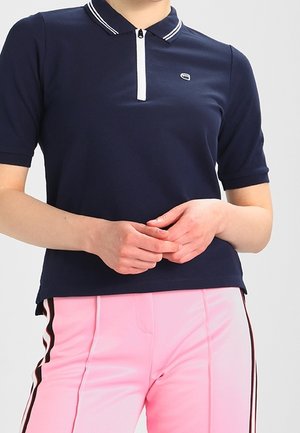 Osoba ubrana w granatową koszulkę polo z krótkim rękawem, z białymi paskami na kołnierzyku oraz różowe spodnie z czarno-białymi paskami po bokach.