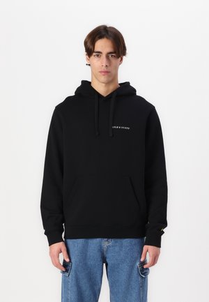 Sort hættetrøje lavet af blødt stof, med en frontlomme, snoretræk og hvidt logo på venstre bryst. Båret med blå jeans.
