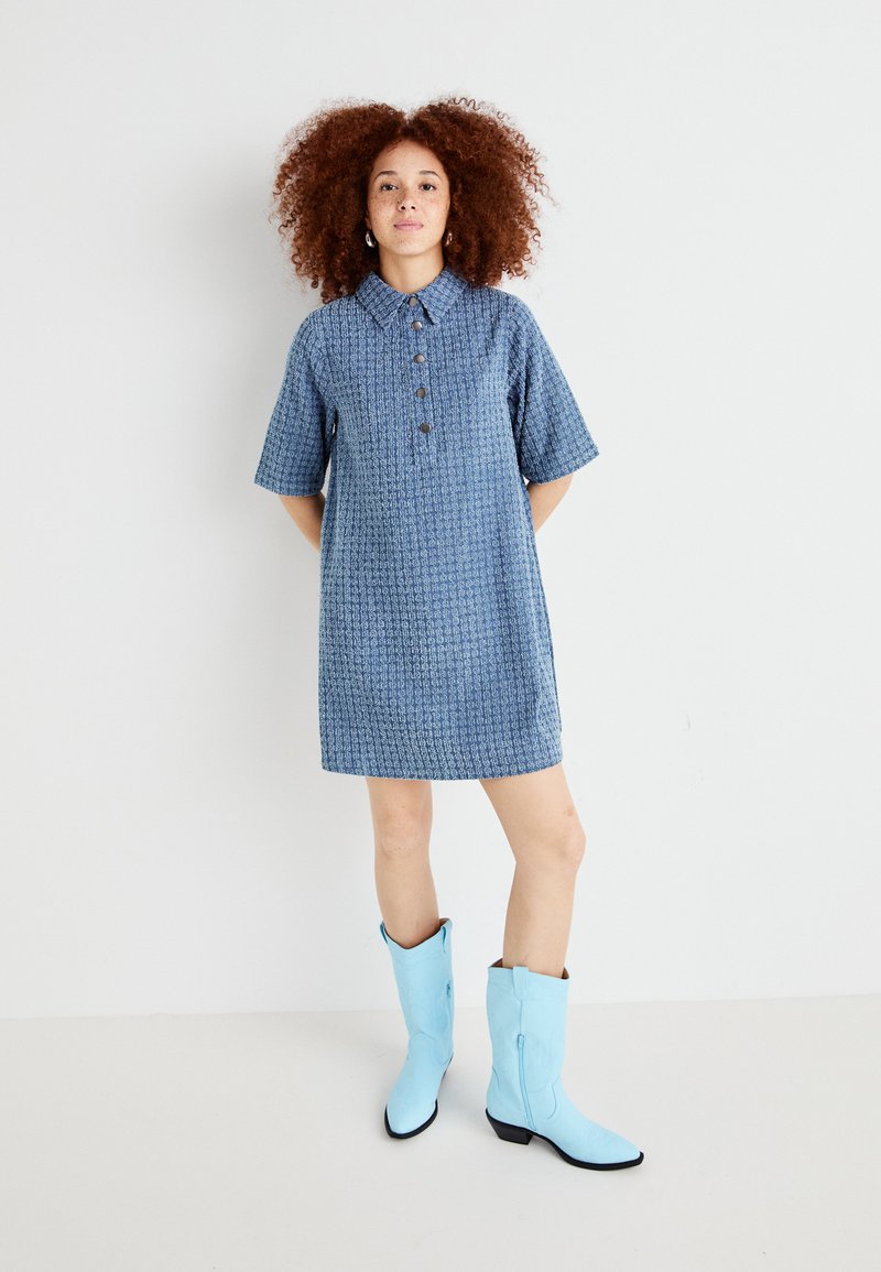 Robe bleue à manches courtes avec un col, présentant un motif texturé et des détails de boutons. Portée avec des bottes de cowboy bleu clair.