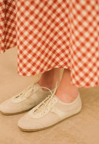 Scarpe basse in suede beige con chiusura lace-up, caratterizzate da bordi scallop e suola in gomma, abbinate a una gonna a quadri rossi e bianchi.