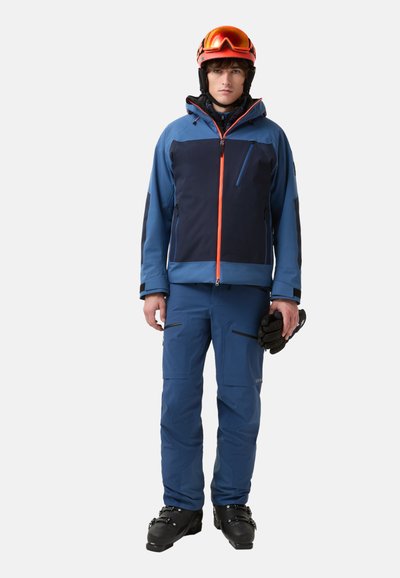 Ski outfit featuring a blue jacket with navy accents and orange zipper, blue pants, and black ski boots, held gloves in hand.