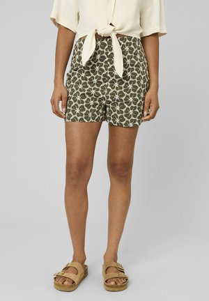 Femme portant un short vert olive avec une broderie florale blanche, associé à une chemise crème à manches courtes nouée et des sandales beige à boucle.