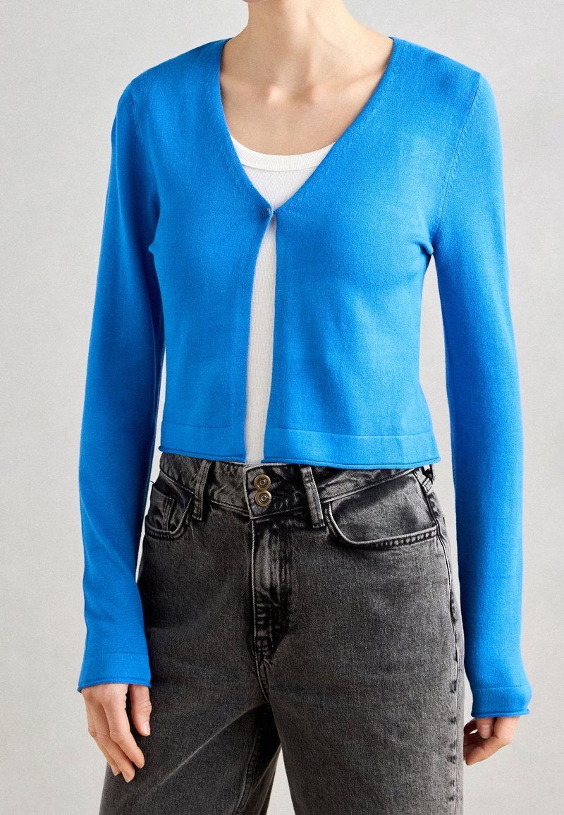 Femme portant un cardigan court bleu vif à col en V par-dessus un haut blanc et un jean denim noir taille haute avec double boutons.