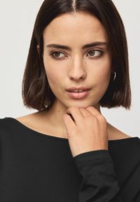 Sort top med bred halsudskæring og lange ærmer. Nærbillede af en model med skulderlangt mørkt hår, let makeup og en hånd på hagen.