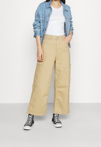 Beige široké cargo kalhoty s bočními kapsami, doplněné bílým topem a světle modrou denimovou bundou. Černé vysoké tenisky doplňují celý outfit.