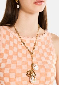 Gul kedjehalsband med stora länkar, prydt med korall- och snäckpendlar. Bärs över en peach-färgad topp med rutmönster och vågiga kanter.