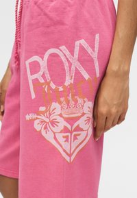 Ροζ σορτς με κορδόνι, με το κείμενο "ROXY", floral σχέδιο και έμβλημα κορώνας κοντά στην πλαϊνή τσέπη, φορεμένα από ένα άτομο.
