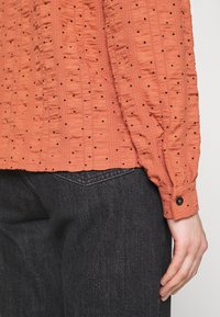 Lys orange tekstureret bluse med små runde udsparinger, sammensnørede ærmer og en lukning med knap, parret med mørke denim jeans.