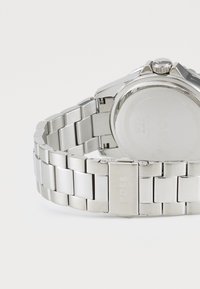 Orologio in acciaio inox con bracciale a maglie, caratterizzato da una finitura liscia, una cassa rotonda e una corona decorativa. Logo inciso sulla chiusura.