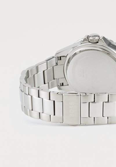 Montre en acier inoxydable avec un bracelet à maillons, dotée d'une finition lisse, d'un boîtier rond et d'une couronne décorative. Logo gravé sur le fermoir.