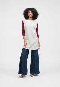 Pelēka bezroku tunika ar apaļu kakla izgriezumu, pārlaista pāri tumši sarkanai garroku kreklai. Kopā valkātas tumši zilas džinsa platās bikses un sandales.