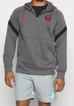 Mand iført en grå Paris Saint-Germain hættetrøje med røde detaljer og lyseblå Nike-shorts står mod en ensfarvet baggrund.