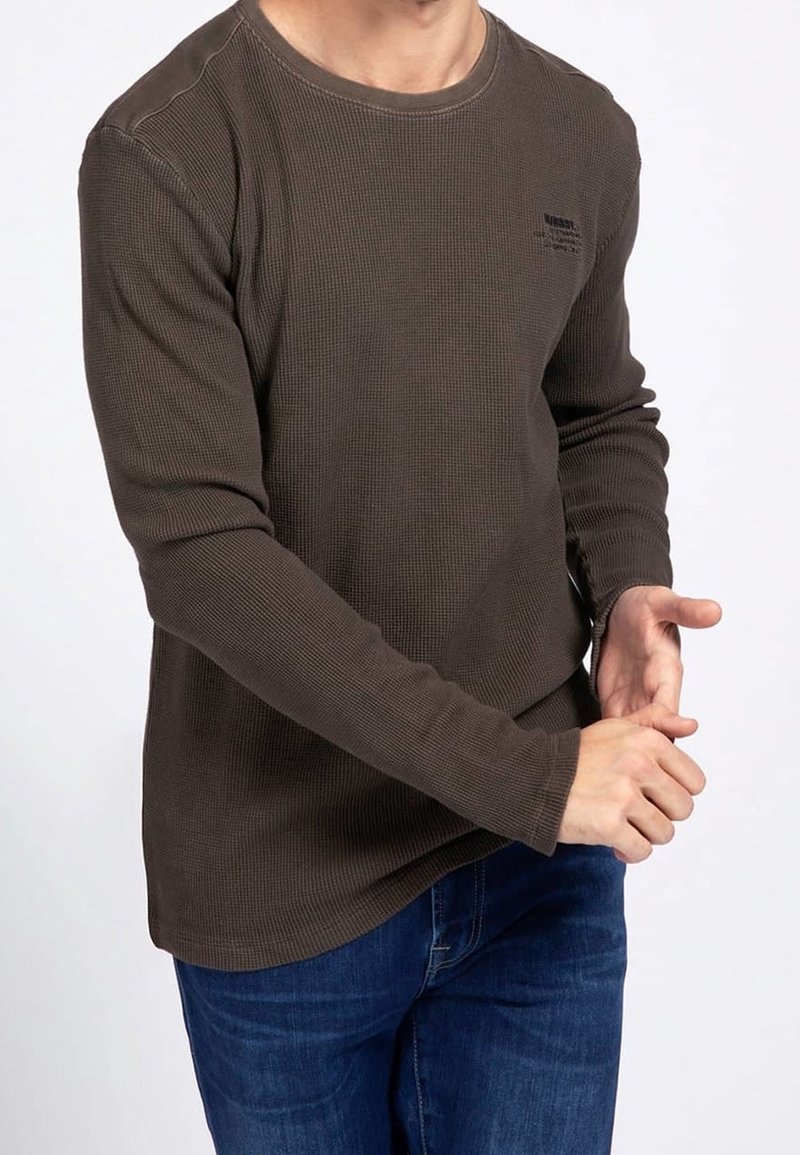 Homme portant une chemise en maille gaufrée marron foncé à manches longues et un jean bleu, ajustant la manche avec ses mains sur un fond uni.