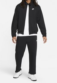 Svart Nike zip-up jacka med krage, parat med svarta sweatpants. Vit Nike-logotyp på jackan och byxorna. Vita sneakers fulländar outfiten.