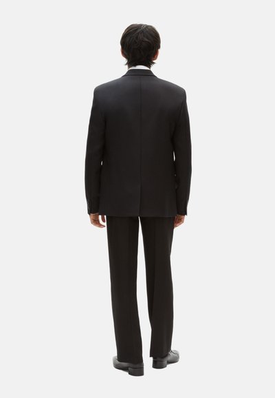 Schwarzer Anzug mit einem taillierten Sakko, das mit einem Revers und einem einzelnen Schlitz versehen ist, kombiniert mit geraden Hosen und schwarzen Schuhen.
