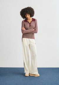 Mulher com cabelo cacheado está em pé num tapete azul, vestindo uma camisola polo de manga comprida cor de vinho, calças brancas de pernas largas e sapatos plataforma, com os braços cruzados.