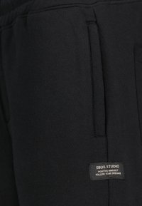 Schwarze Baumwoll-Jogginghosen mit seitlichen Taschen. besitzt ein genähtes Etikett mit der Aufschrift "SBLV STUDIO" sowie motivierendem Text in hellem Schriftbild. Weiche Textur.