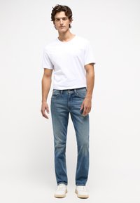 Vit crew-neck t-shirt, blå urtvättade jeans, ljusa sneakers. Enkelt design, klassisk passform, med synlig sömnad och lätt slitage på jeans.