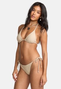 Beżowy bikini z prążkowanej tkaniny, z trójkątnym stanikiem i regulowanymi trokami po bokach majtek. Uzupełnione srebrnym naszyjnikiem łańcuszkowym.