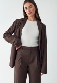 Hnedý tailored blejzer na bielom rebrovanom svetri, doplnený zodpovedajúcimi hnedými nohavicami. Obsahuje ostré klopy a štíhly strih.