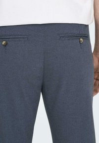 Pantalones texturizados azul marino con dos bolsillos traseros, con botones de latón y un corte recto. El material parece tejido con un patrón sutil.