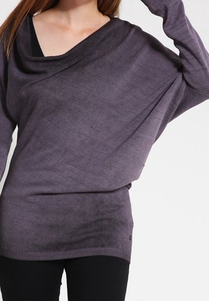 Vrouw draagt een loszittende, donkergrijze trui met lange mouwen over een zwart topje, met één arm omhoog en zwarte broek.