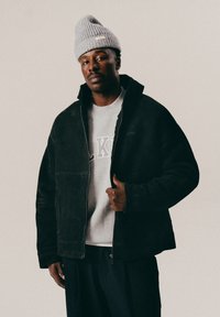 Man wearing a gray knit beanie, black jacket, and light gray sweatshirt with text, standing against a plain light background.