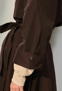 Brun ydertøj med en glat tekstur, udstyret med et bælte i taljen og knapdetaljer ved ærmerne, lagdelt over en beige ærme.