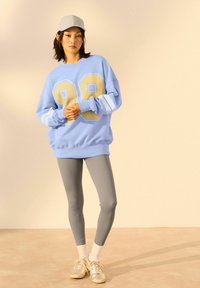 Felpa azzurra con grande logo giallo "99", strisce bianche sulle maniche, abbinata a leggings grigi a coste e sneakers argentate. Cappellino grigio.